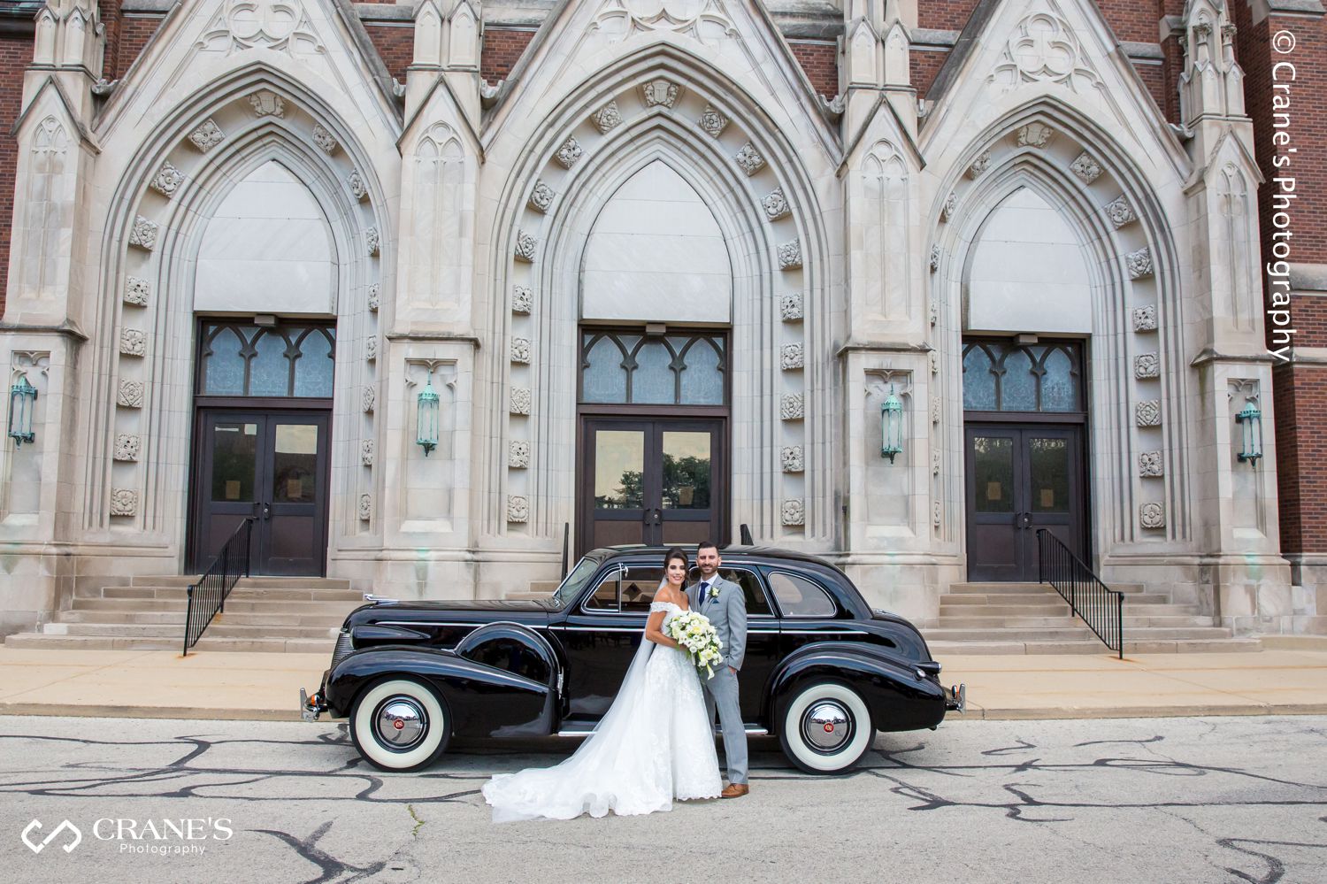 A portrait of a bride and groom with a vintage car in front of St. Peter and Paul Church in Naperville