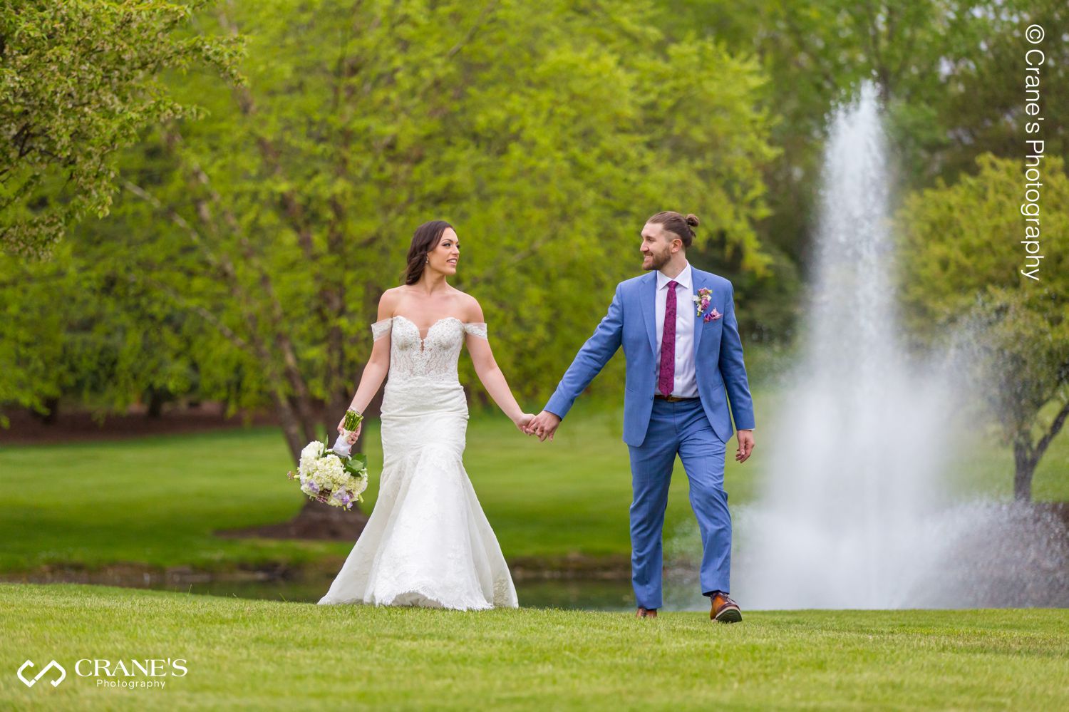 A photo of a bride and groom walking hand in hand with a beautiful fountain in the background
