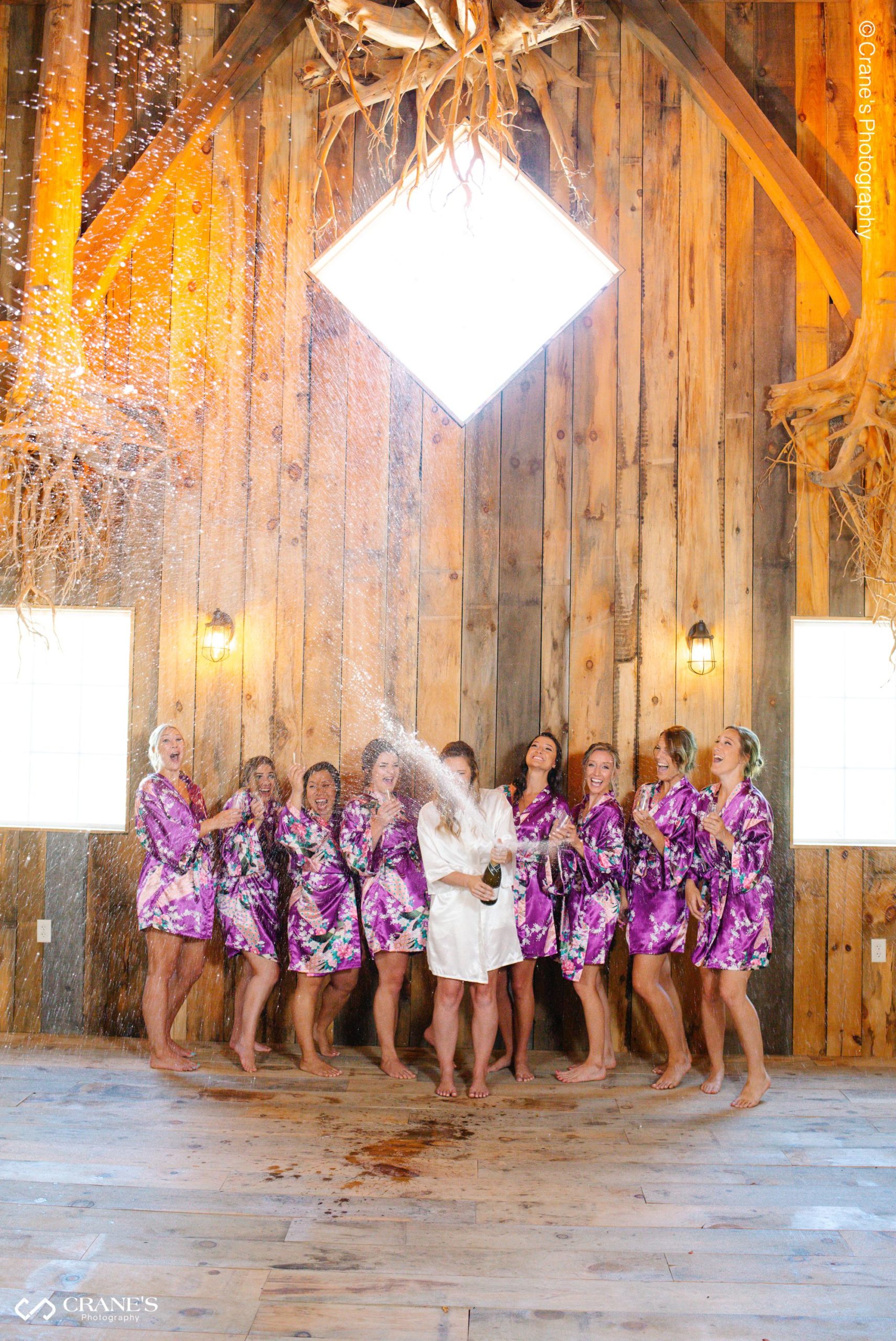 A bride is popping a champagne with her bridesmaids on her wedding day at The Swan Barn Door