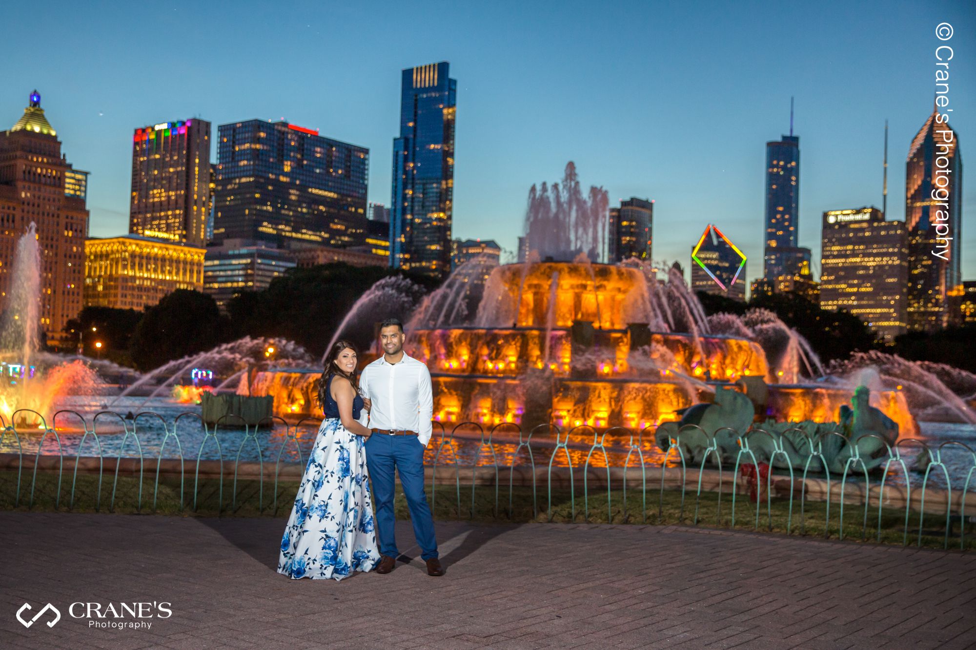 A couple pose in front of the Buckingham Fountain in Chicago with the skyline at night in the background