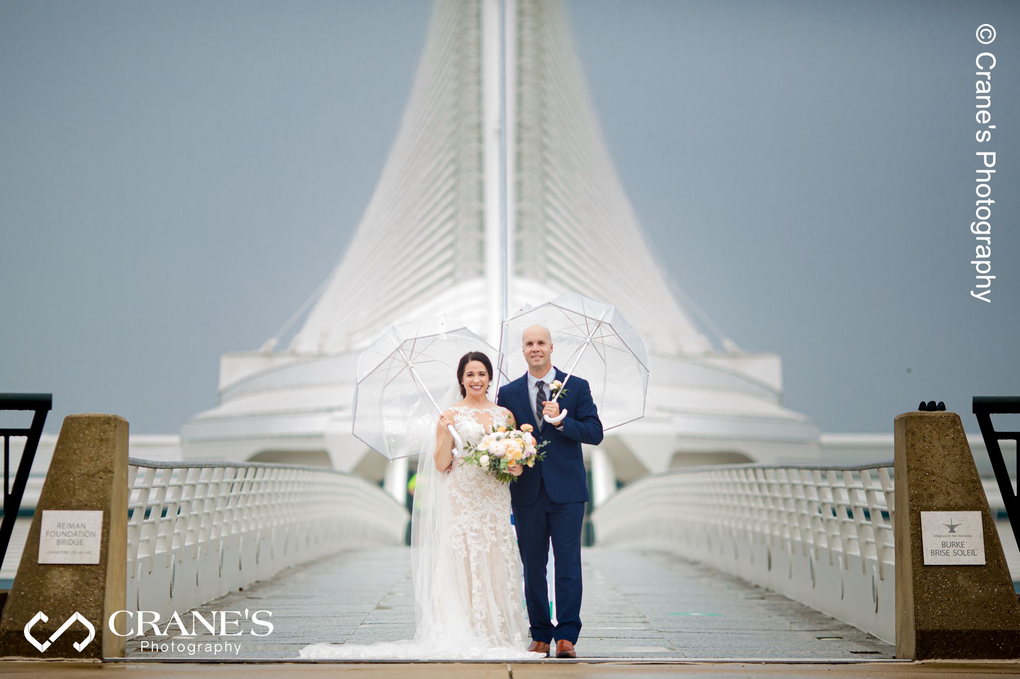 Bride and groom holding clear umbrellas posing for a wedding photo at the bridge in front of Art Museum in Downtown Milwaukee