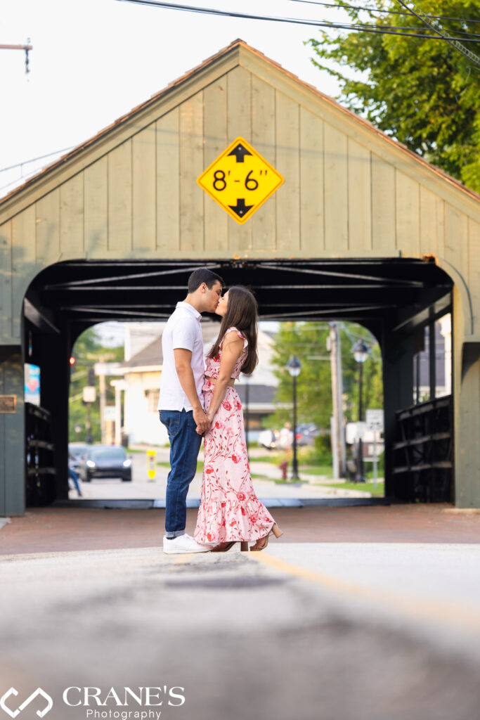 An engaged couple are kissing in downtown Long Grove with the bridge in the background.