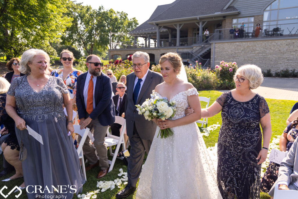 A bride is walking down the aisle escorted by her parents outside Bloomingdale Golf Club.