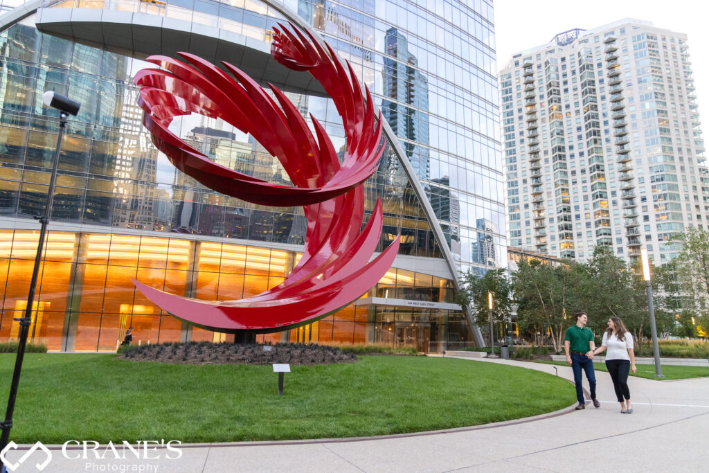 An engaged couple are posing for a photo by the Constellation sculpture at the Riverwalk in Chicago.
