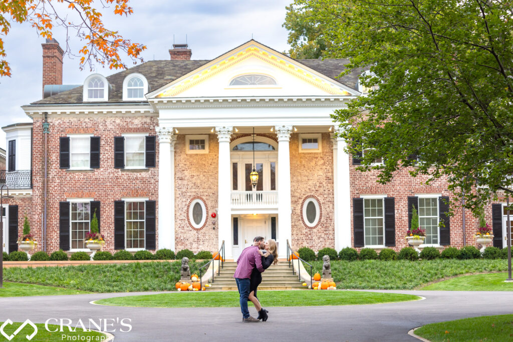A couple sharing a romantic kiss with the Robert R. McCormick House in the background, set against the stunning fall landscape of Cantigny Park.