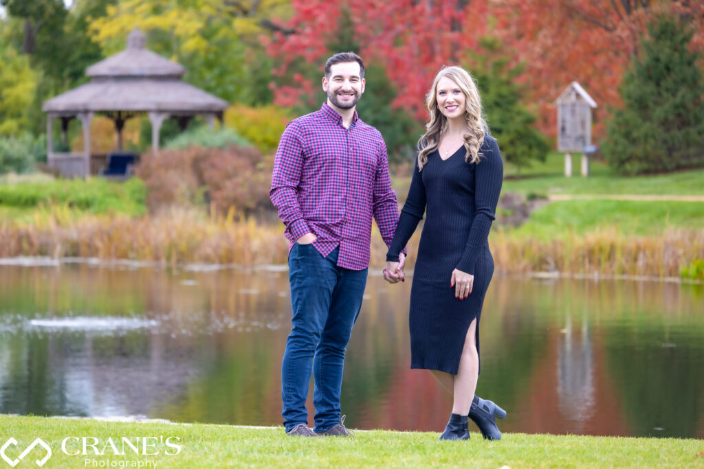 An engaged couple dressed casually, posing for an engagement photo by the pond at Cantigny Park, with a stunning red tree in the background.