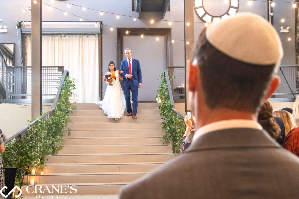 A heartwarming photo during a Jewish wedding ceremony of a bride and her dad walking down the stairs at Artifact Events, with the groom in the foreground.