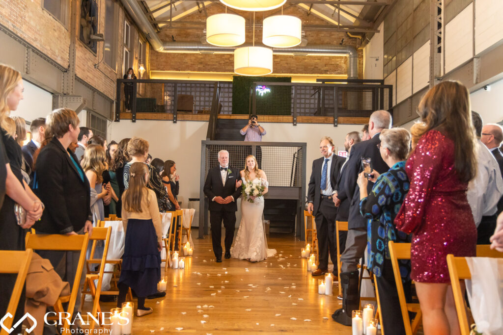 Captured at City View Loft, a beautiful scene unfolds as a bride, adorned in an elegant wedding gown, walks down the aisle accompanied by her father, both radiating joy during their heartfelt wedding ceremony.