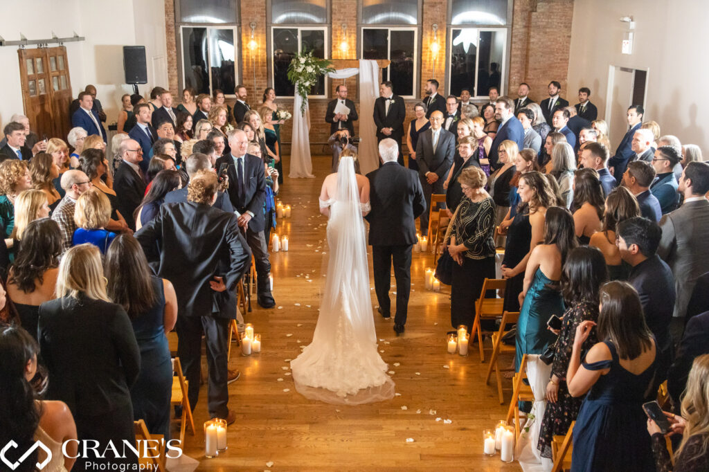 A heartwarming moment captured at City View Loft, where a bride, dressed in a stunning wedding gown, walks down the aisle with her proud father by her side during their wedding ceremony.