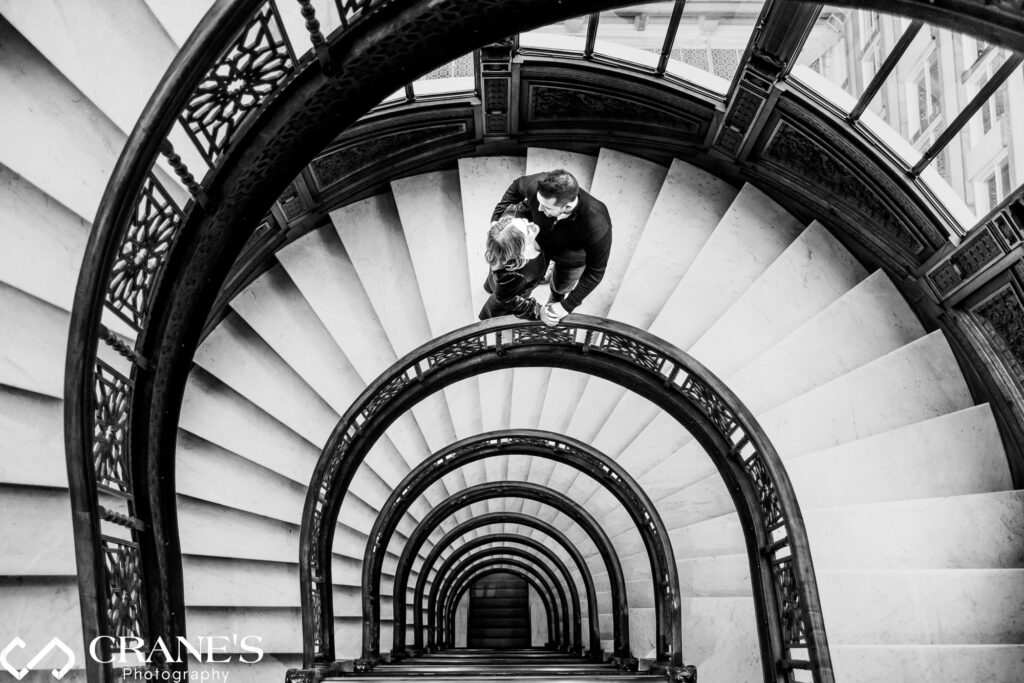 An elegant black and white portrait capturing the love between an engaged couple, framed against the elegant backdrop of the spiral staircase at the historic Rookery Building in Chicago.