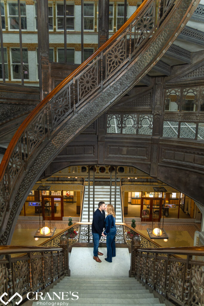 Capturing a tender moment, an engaged couple shares a heartfelt kiss on the iconic spiral staircase at the Rookery Building, Chicago, surrounded by the timeless beauty of their love and the architectural splendor of the location.