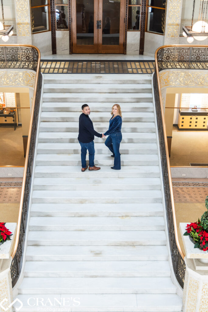 An engagement session photo of a couple gracefully ascends the grand staircase in the Light Court at the Rookery Building in Chicago, their journey symbolizing love and commitment amidst the historic ambiance of this iconic architectural masterpiece.