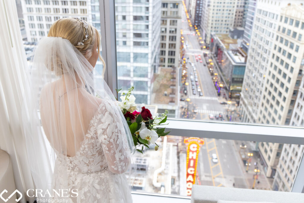 Radiant bride, adorned with a tiara and wedding veil, poses for a photo in the luxurious theWit hotel suite. The backdrop showcases a magnificent view of the city, featuring the iconic 'Chicago' sign at the Chicago Theatre, adding a touch of urban charm to this enchanting bridal moment.