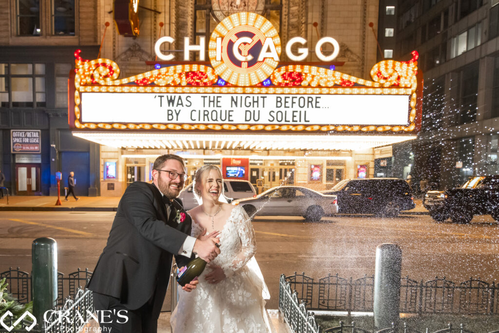 In a festive celebration, the bride and groom joyfully spray champagne at the conclusion of their wedding at theWit, with the iconic Chicago Theatre illuminated in the background.