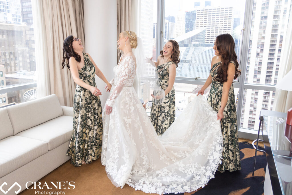 Bridesmaids share laughter and joy while assisting the bride with her wedding gown in a theWit hotel suite, featuring a stunning city view as the backdrop.