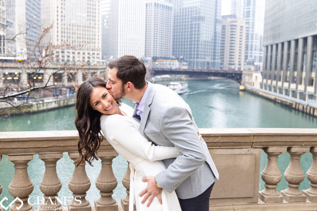 Caught in a joyful moment during their winter engagement session in Downtown Chicago, the guy plants a sweet kiss on his girl's cheek, capturing the warmth of their love amidst the chilly urban backdrop.