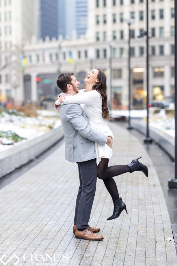 In the wintery ambiance of downtown Chicago, an engaged couple enjoys a moment of joy as the guy lifts his girl, both sharing laughter during their enchanting engagement photoshoot.