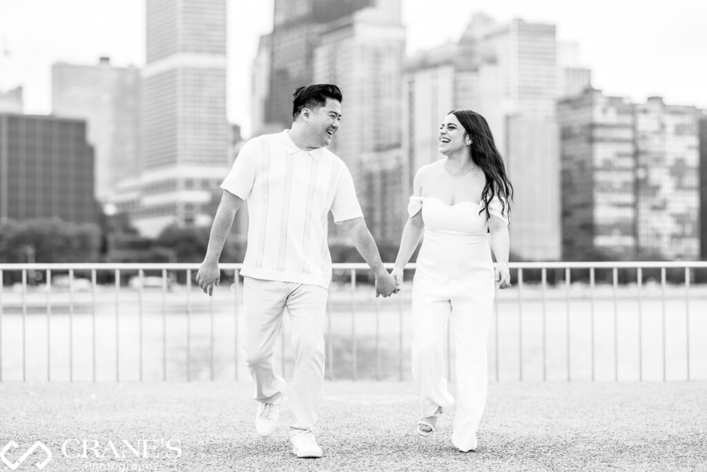 A candid engagement photo of a couple walking together at Olive Park in Chicago. They hold hands and smile at each other, with the city's skyline in the background. The scene captures a moment of joy and connection between them.