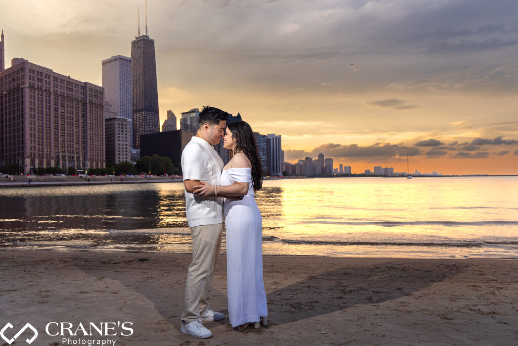 A happy engaged couple pose for a photo at Olive Park, with a spectacular sunset and the downtown skyline in the background. The sky is a blend of warm colors, highlighting the city's tall buildings and creating a romantic atmosphere.