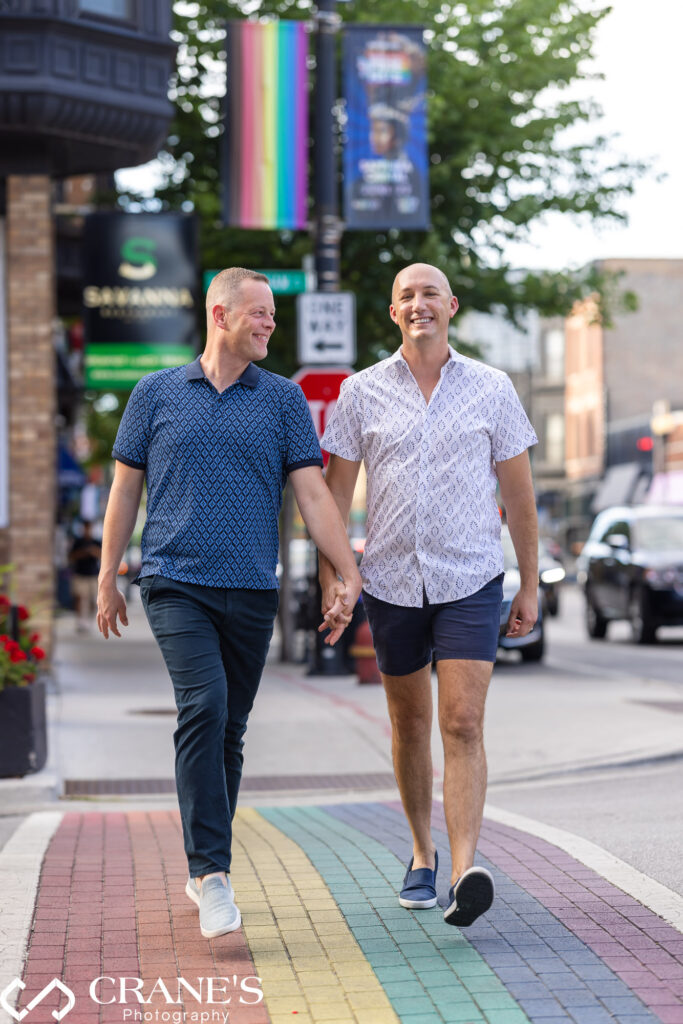 Celebrating their LGBTQ+ engagement in Chicago's North Halsted neighborhood, a gay couple walks hand-in-hand across a colorful rainbow crosswalk.