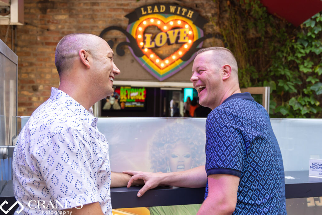 A joyous gay couple, celebrating in a North Halsted bar in Chicago with a vibrant LGBTQ+ pride flag hanging proudly behind them.