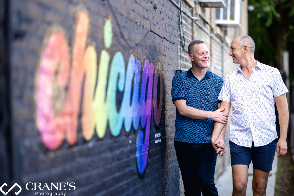 A LGBTQ+ couple stroll hand-in-hand through North Halsted with a vibrant street art depicting Chicago in rainbow colors brightening the scene next to them.