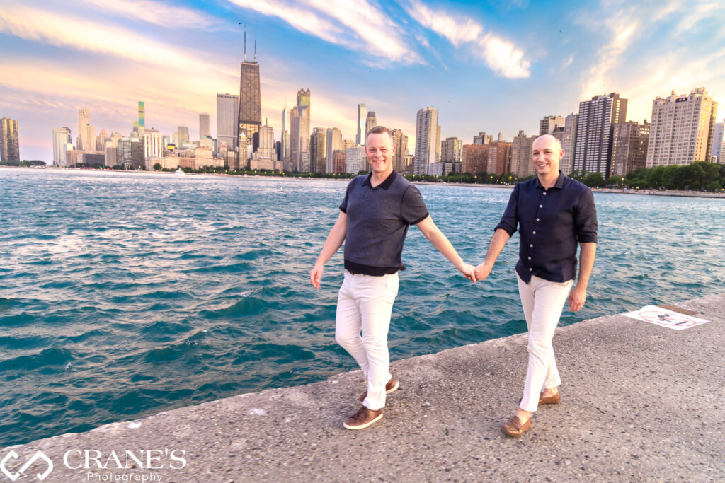 Two people, dressed for a celebration, walk hand-in-hand along North Avenue Beach in Chicago. The sky is ablaze with orange and pink hues of sunset, casting a warm glow on the couple and the city skyline in the distance. (LGBTQ+ engagement)