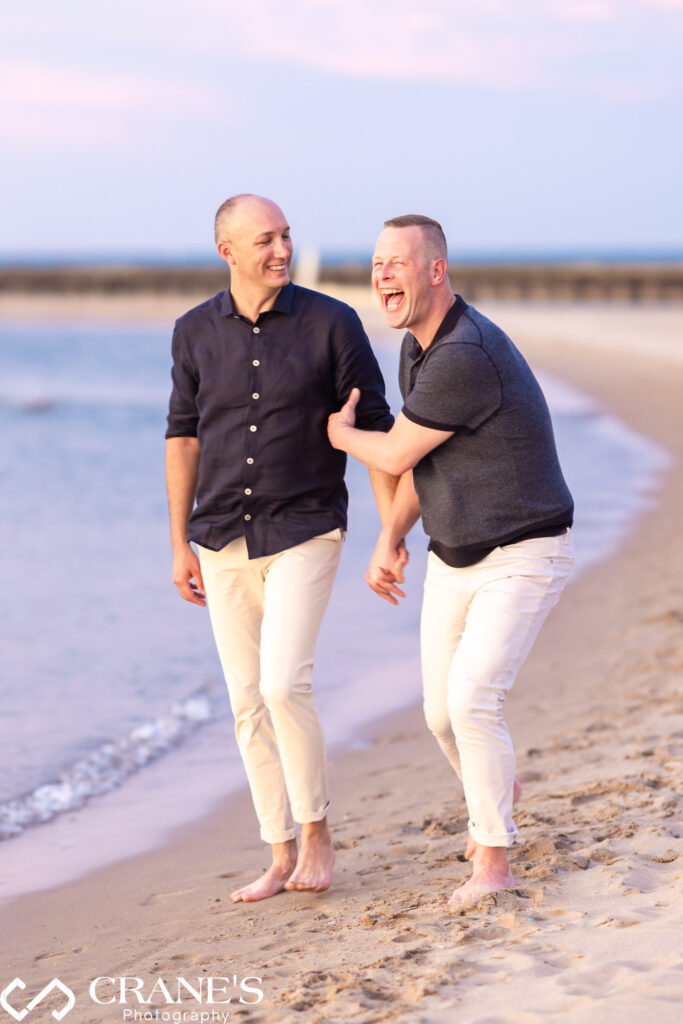 Two men, celebrating their LGBTQ+ engagement, walk hand-in-hand along North Ave Beach in Chicago with pants rolled up, enjoying a carefree moment.