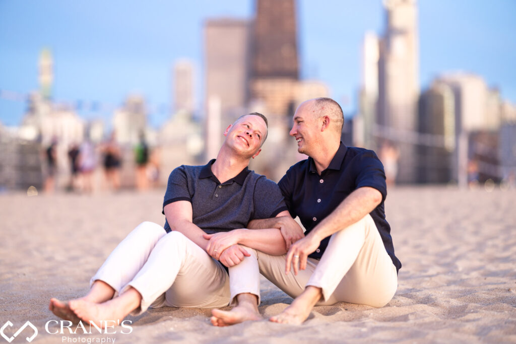 A LGBTQ+ couple, celebrating their engagement on Chicago's North Avenue Beach, sit nestled in the golden sand as the sun dips behind the city skyline, painting the sky with vibrant hues.