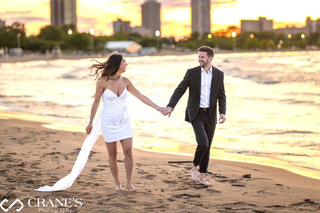 An engaged couple are walking on North Ave Beach with stunning sunset behind them.