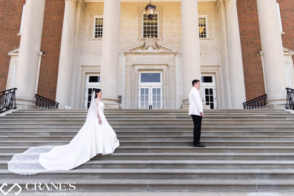 A bride and groom share a heartfelt first look moment on the grand staircase at the Chicago History Museum. The bride, wearing a flowing white gown, and the groom, dressed in a sharp white suit, exchange joyful expressions amidst the elegant backdrop of the historic venue.