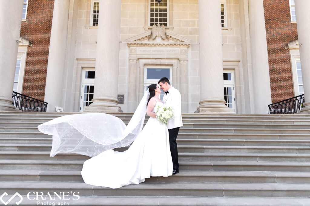 A bride and groom kiss on their wedding day on the grand staircase at the Chicago History Museum. The wind picks up the bride's veil, creating a dramatic and romantic moment against the elegant backdrop of the historic venue.