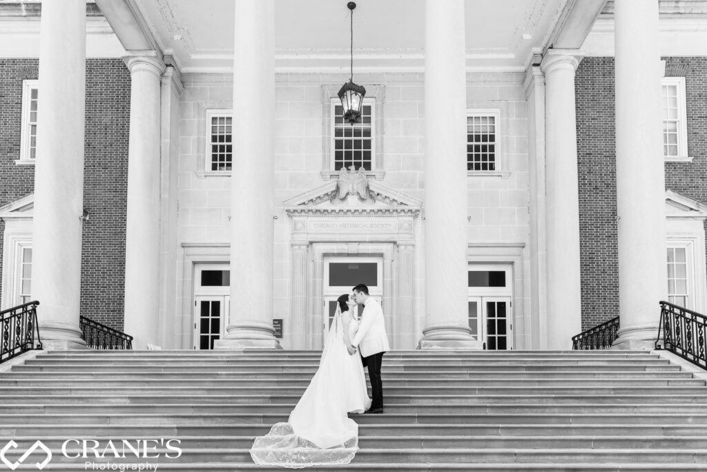 Newlyweds in a tender moment at the Chicago History Museum. Groom kisses bride's lips, her train cascading down the grand staircase. Timeless wedding portrait with historic backdrop.