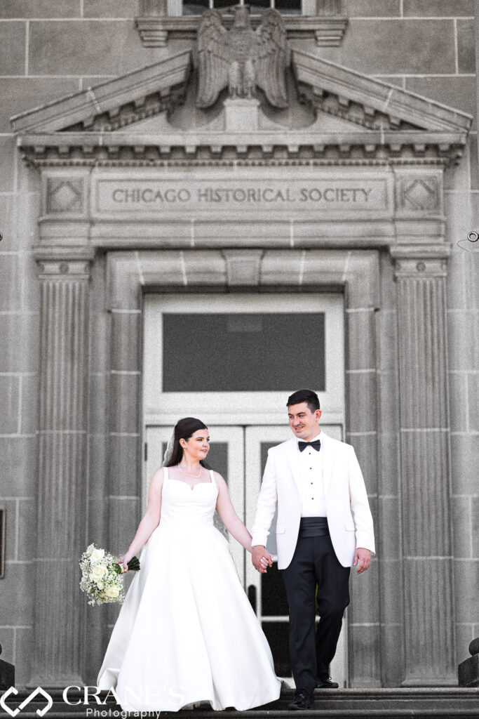 A dramatic, award-winning wedding photo of a couple walking down the grand staircase at the Chicago History Museum. The bride's flowing gown and the groom's stylish suit are highlighted against the Chicago Historical Society sign at the venue.