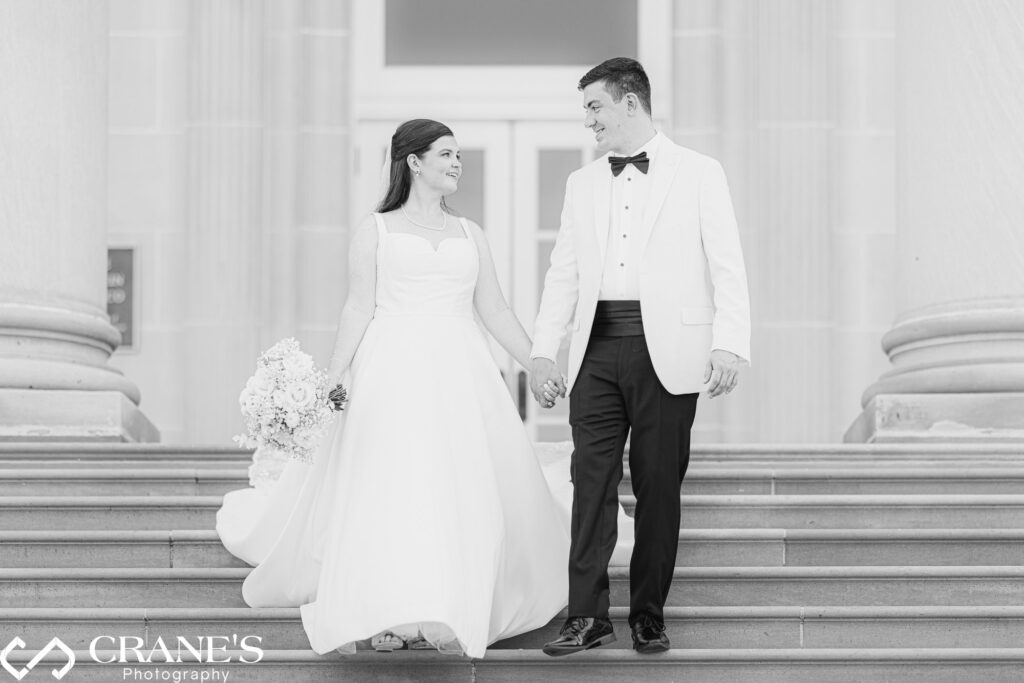 An elegant wedding photo of a bride in a flowing white dress and a groom in a white suit walking down the grand staircase at the Chicago History Museum. The historic venue provides a sophisticated backdrop for the couple's special moment.