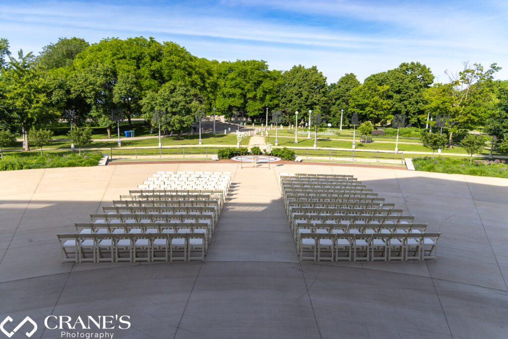 Wedding ceremony at Uihlein Plaza, Chicago History Museum. White chairs line the aisle. Lush greenery of Lincoln Park and flower arch form a picturesque backdrop.