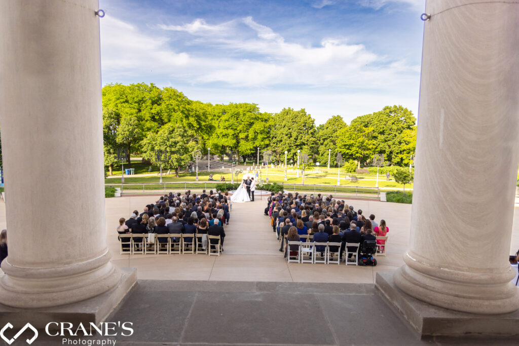 An outdoor wedding ceremony at the Chicago History Museum, with blue skies and the lush greenery of Lincoln Park in the background. A flower arch adds to the beauty of the special occasion.