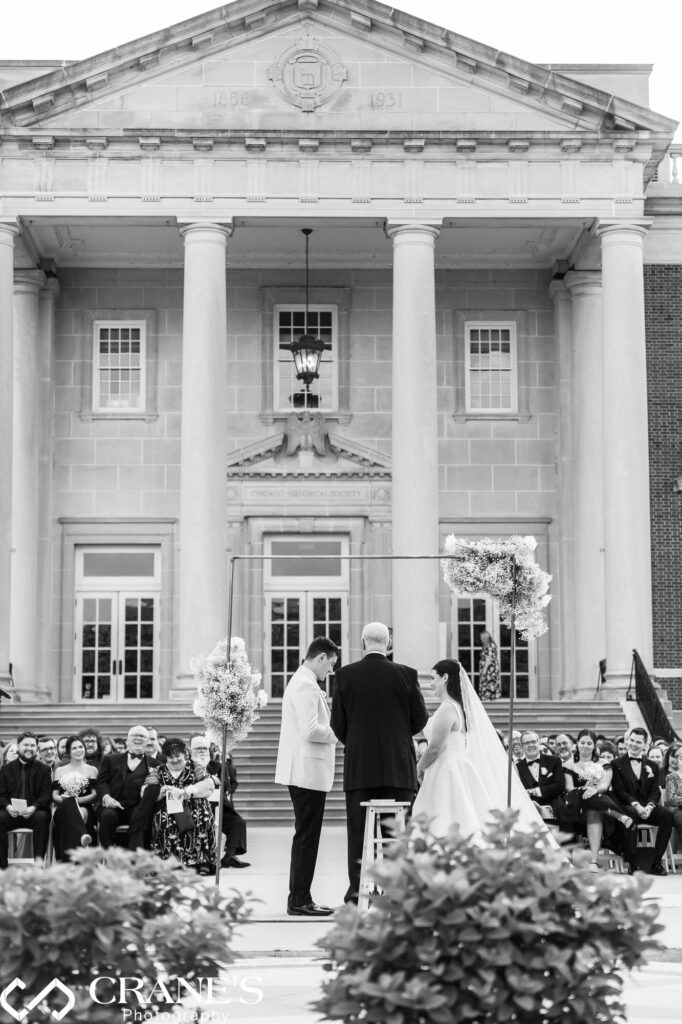 The bride and groom exchange vows during an outdoor wedding ceremony, with the grand architecture of the Chicago History Museum in the background. The elegant venue adds a historic and majestic touch to the couple's special moment.