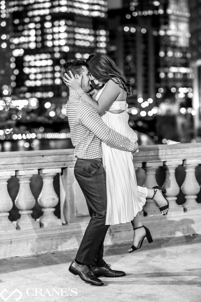 Black and white engagement photo in Chicago of a joyful couple at nighttime. The woman is lifted in the air, playfully kissing the man.