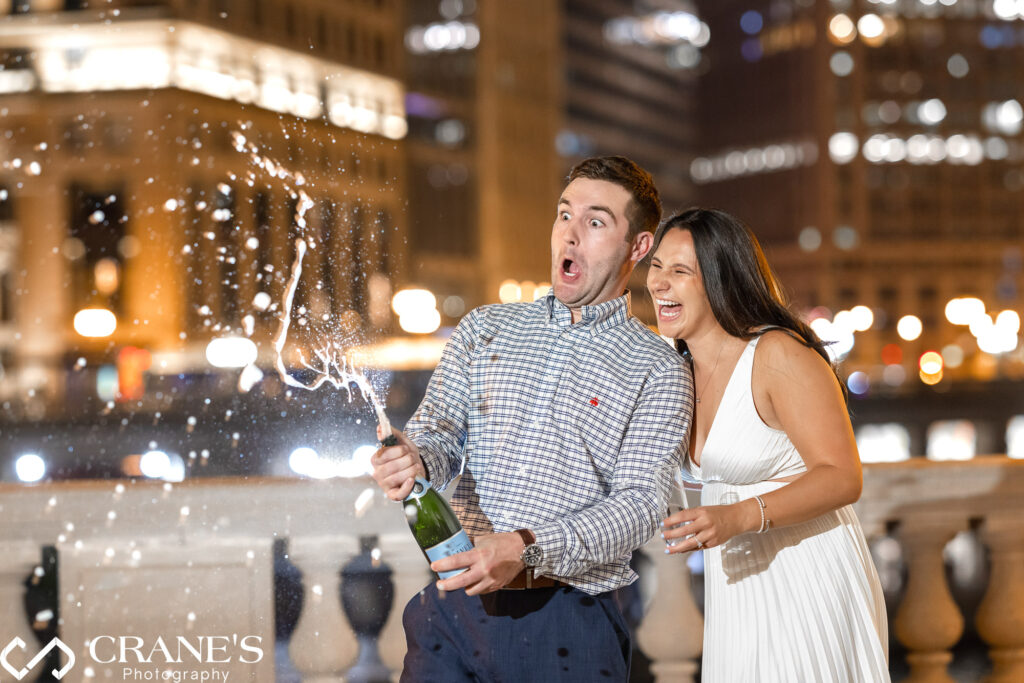 A night-time Chicago engagement photo captures a celebratory toast. The couple, smiles on their faces, pop a bottle of champagne in front of the iconic Wrigley Building, bathed in warm light.