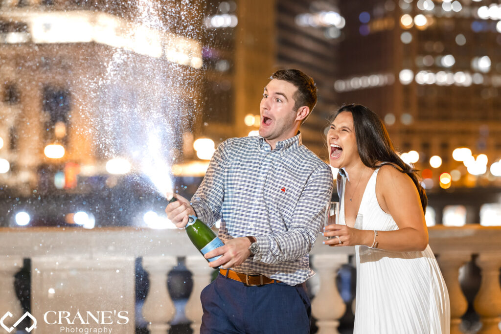 A nighttime engagement session at the Wrigley Building in Chicago, capturing a candid moment of a couple popping champagne."