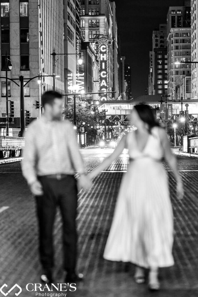 Engaged couple walks hand-in-hand on a Chicago street at night, with the iconic Chicago Theatre sign in focus illuminated behind them.