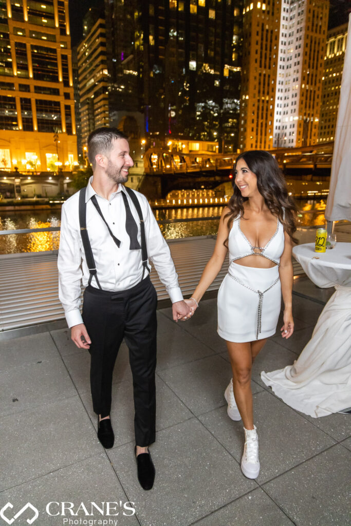 The groom, with his tie loosened, and the bride in a chic white dress pose for nighttime wedding photo on the terrace at RPM Events, with the city river shimmering in the background.