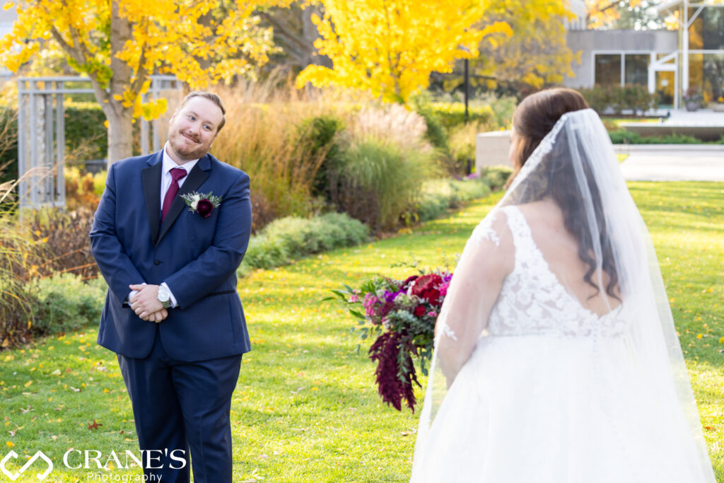 Groom smiling warmly at his bride during their first look at the Morton Arboretum, with vibrant fall foliage and golden trees in the background. The bride, wearing a lace wedding gown with a veil, stands facing him, holding a bouquet of deep red and purple flowers.