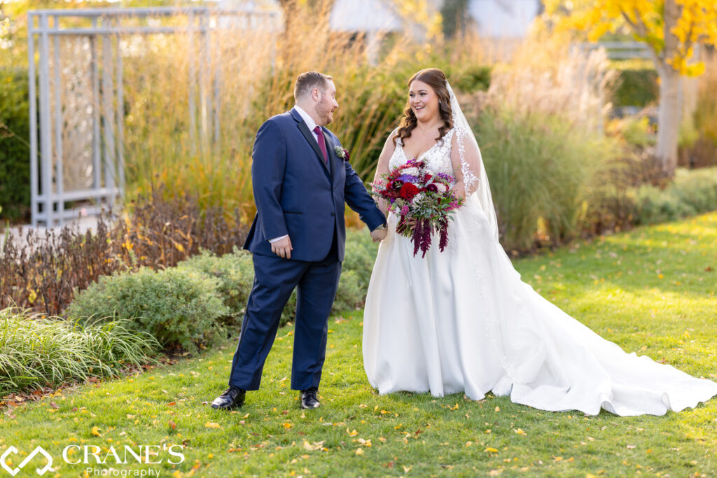 Bride and groom holding hands and smiling at each other as they walk outdoors at the Morton Arboretum, surrounded by the beauty of nature and vibrant fall foliage.