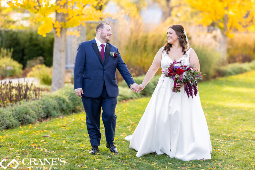 Bride and groom, Julia and David, standing hand-in-hand in front of vibrant autumn foliage at Morton Arboretum.