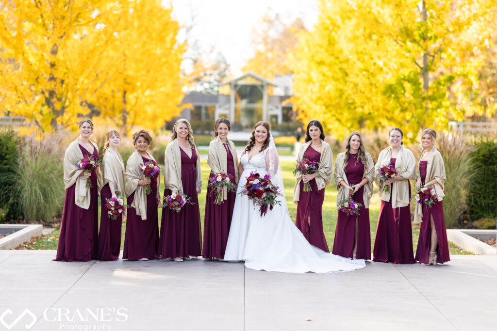 Bridesmaids wearing maroon dresses and beige shawls, standing together in a picturesque outdoor setting at the Morton Arboretum, with soft fall light illuminating the scene.