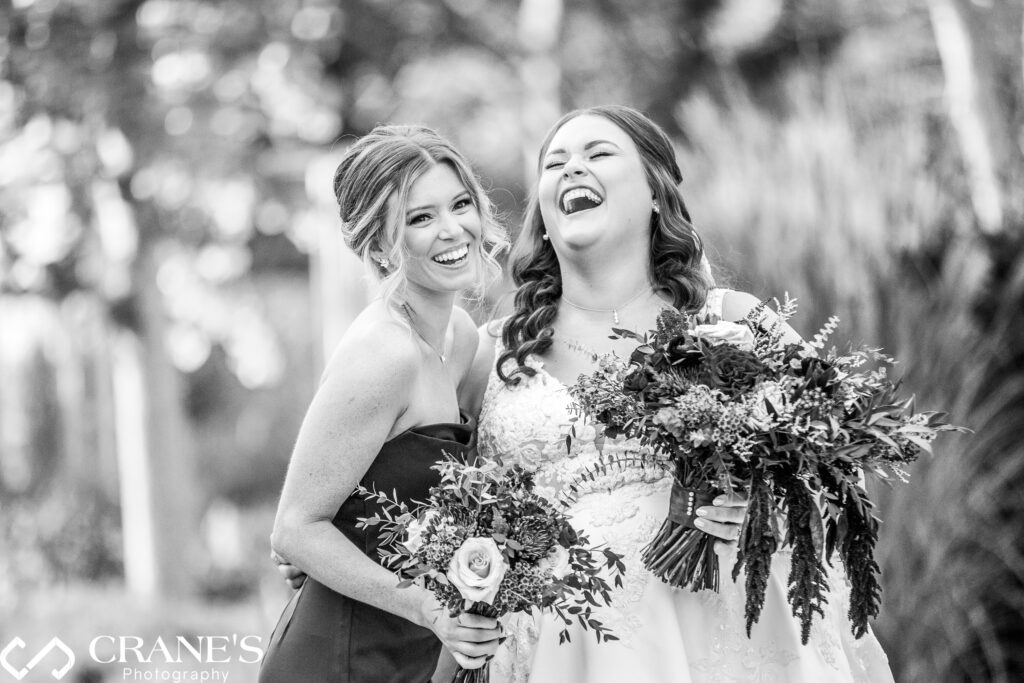 Black and white image of a bride laughing with her bridesmaid at their wedding at the Morton Arboretum. The two share a joyful moment, with the bride holding her bouquet, as the timeless monochrome tones highlight their expressions and the elegant details of the scene.