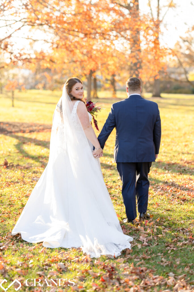 Bride and groom walking hand in hand through a field of fallen autumn leaves at the Morton Arboretum. The bride, glancing back over her shoulder, holds a bouquet of burgundy and purple flowers, while the groom, in a navy suit, walks beside her. The trees behind them are filled with golden leaves, adding a warm glow to the scene.