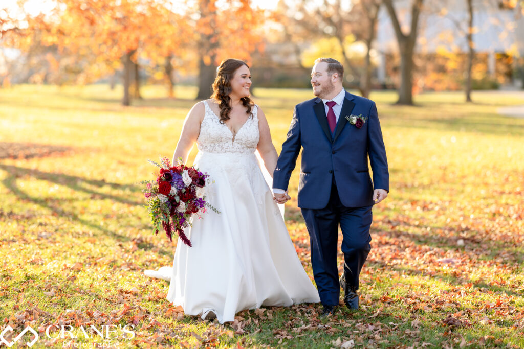 Bride and groom walking hand in hand through a field of fallen autumn leaves, with the warm glow of the setting sun casting a romantic light behind them.